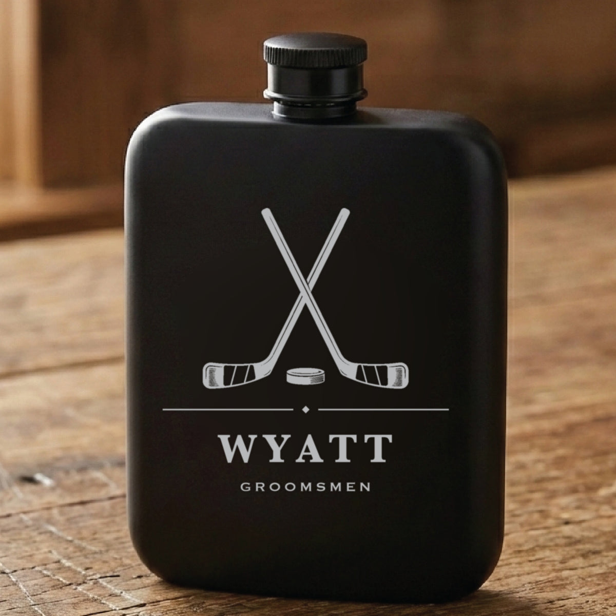 Matte black hockey groomsmen flask laser engraved with crossed hockey sticks and a puck, the name Wyatt and the word Groomsmen.