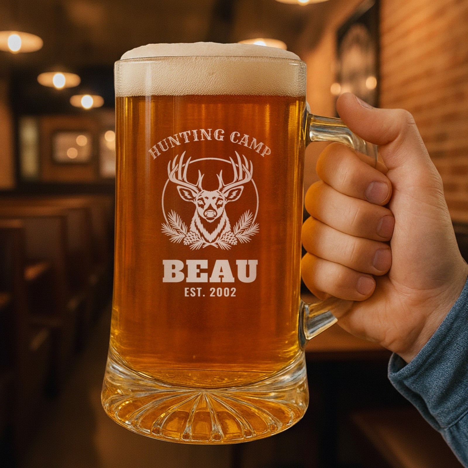 Hand holding the 25 oz personalized hunting camp beer stein in a pub, engraved with a whitetail buck and pine boughs: Hunting Camp, Beau, Est. 2002