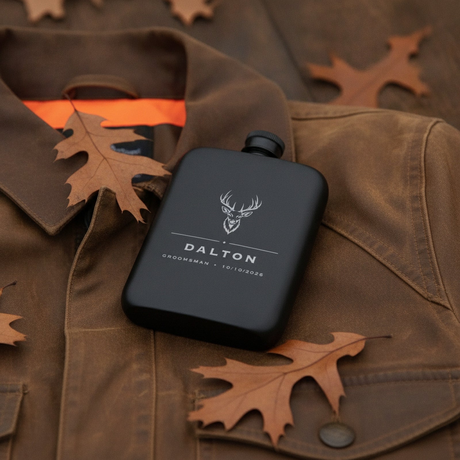 Personalized buck engraved flask resting on a waxed canvas hunting jacket with blaze orange and fallen oak leaves.