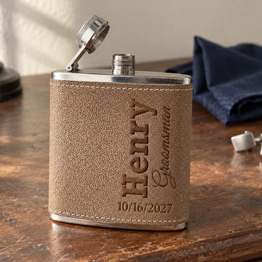 A personalized stainless steel flask with a tan textured leather cover, engraved with the name 'Henry Groomsman' and a date, sitting on a wooden table.