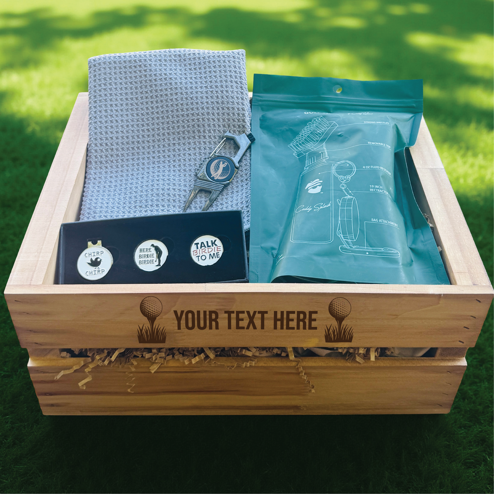 Wooden crate with personalized golf ball markers, gray golf towel, metal divot tool, and turquoise accessories pouch displayed on grass