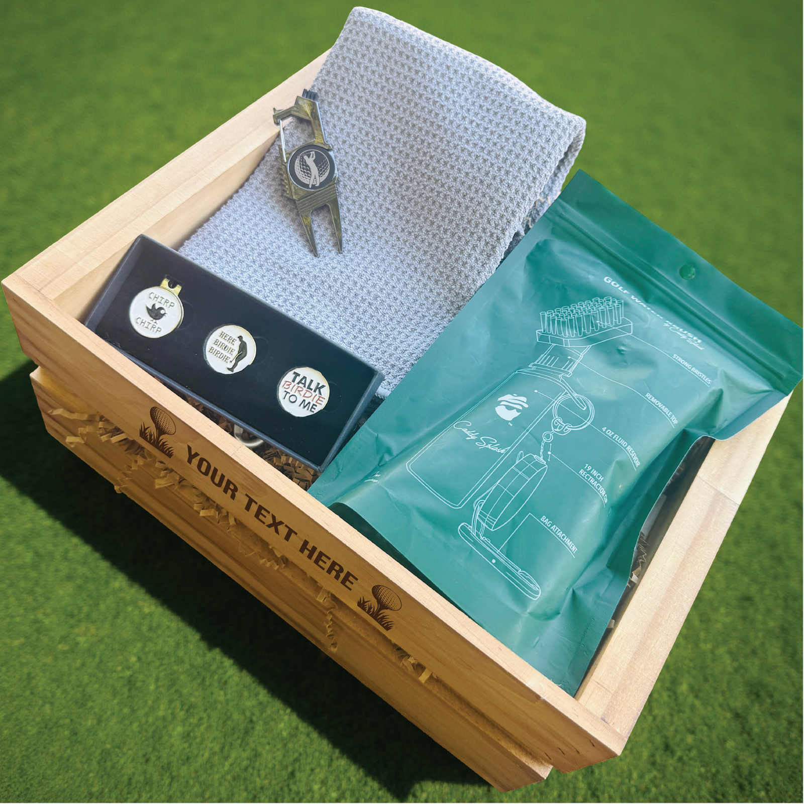 Wooden crate with personalized engraving containing golf towel, divot tool with sample monogram, golf ball markers on black card, and teal accessory pouch on green grass background