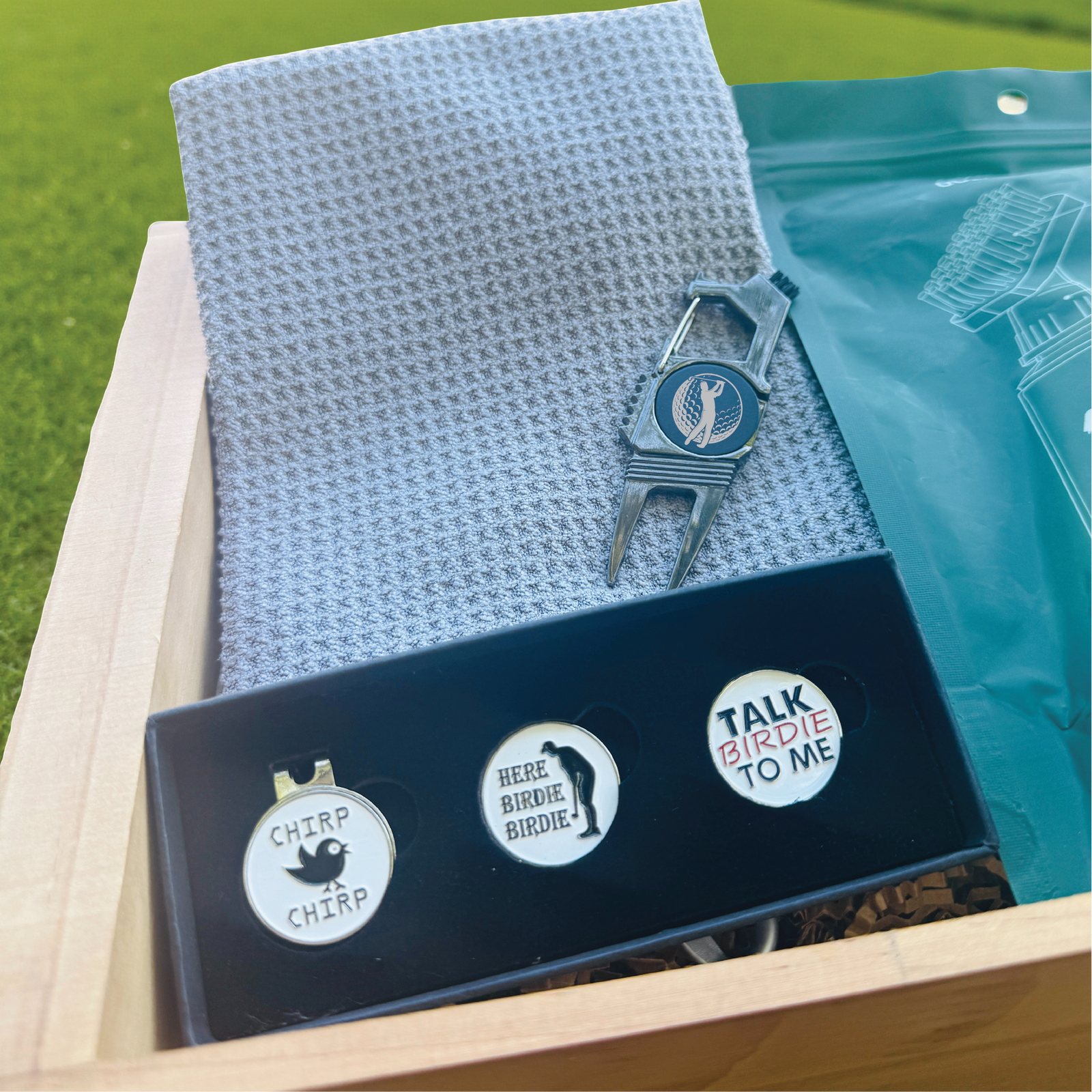 Golf-themed divot tool set with three enamel markers labeled "Chirp Chirp," "Here Birdie Birdie," and "Talk Birdie to Me," displayed in black tray with gray golf towel and silver divot tool in white wooden crate.