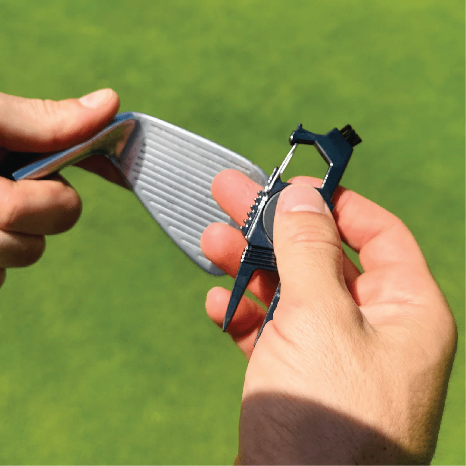 Hands holding a golf ball marker and divot repair tool in black metal against a blurred green golf course background