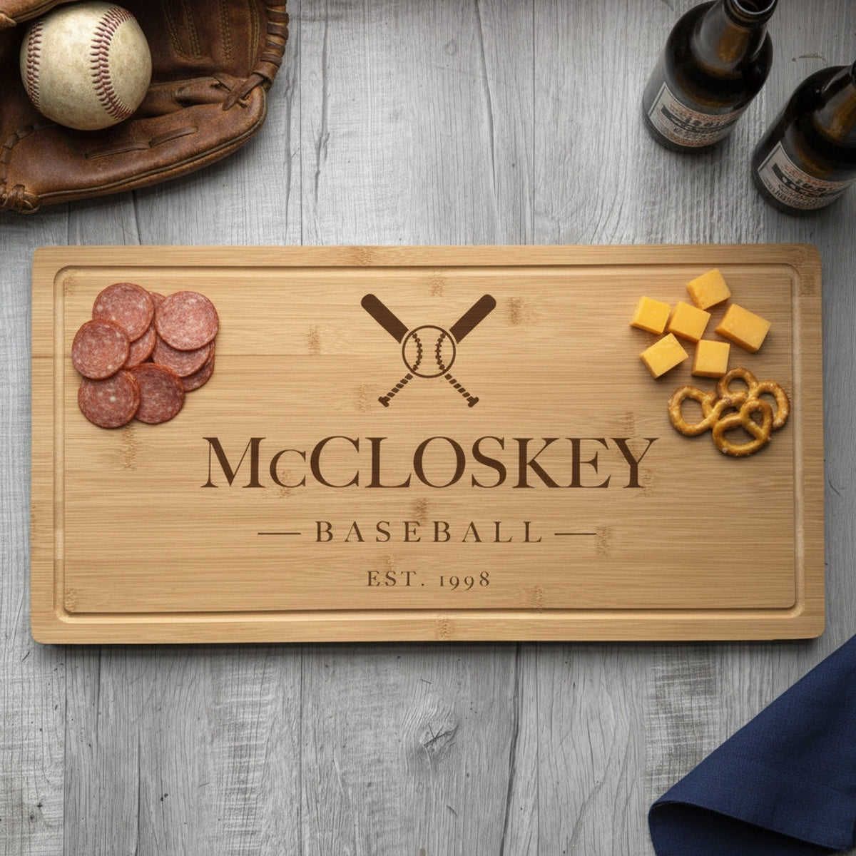 Engraved baseball bamboo board on weathered wood with sliced summer sausage at one end, cheddar and pretzels at the other, beside a glove and two beers.