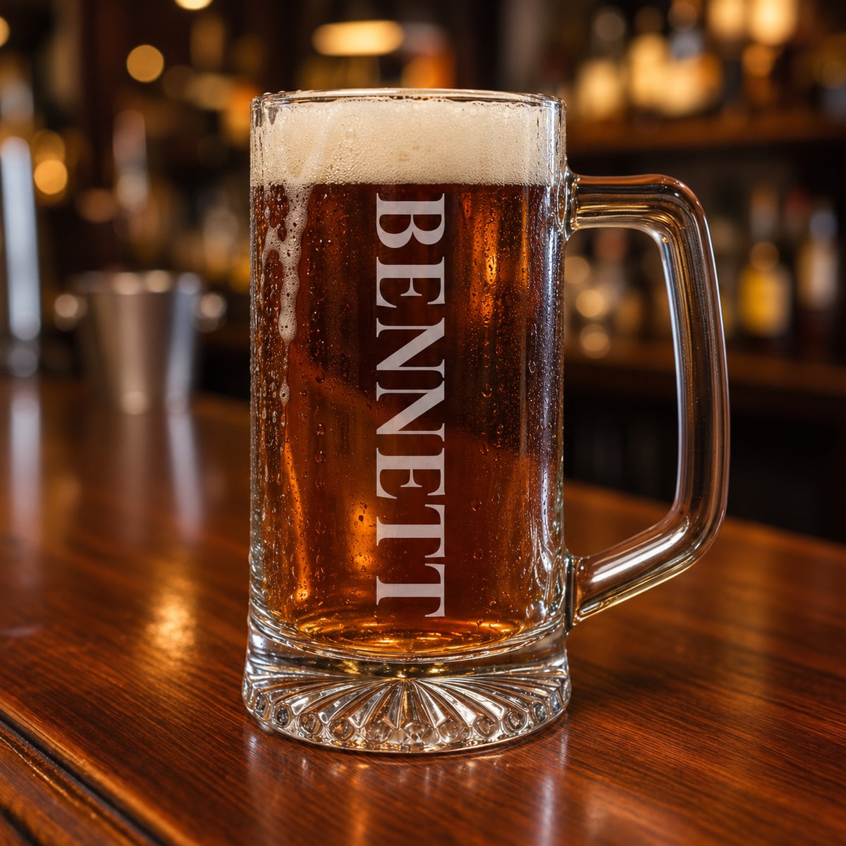 Personalized beer stein full of amber ale on a walnut bar, the name BENNETT engraved in tall frosted letters running down the side of the glass