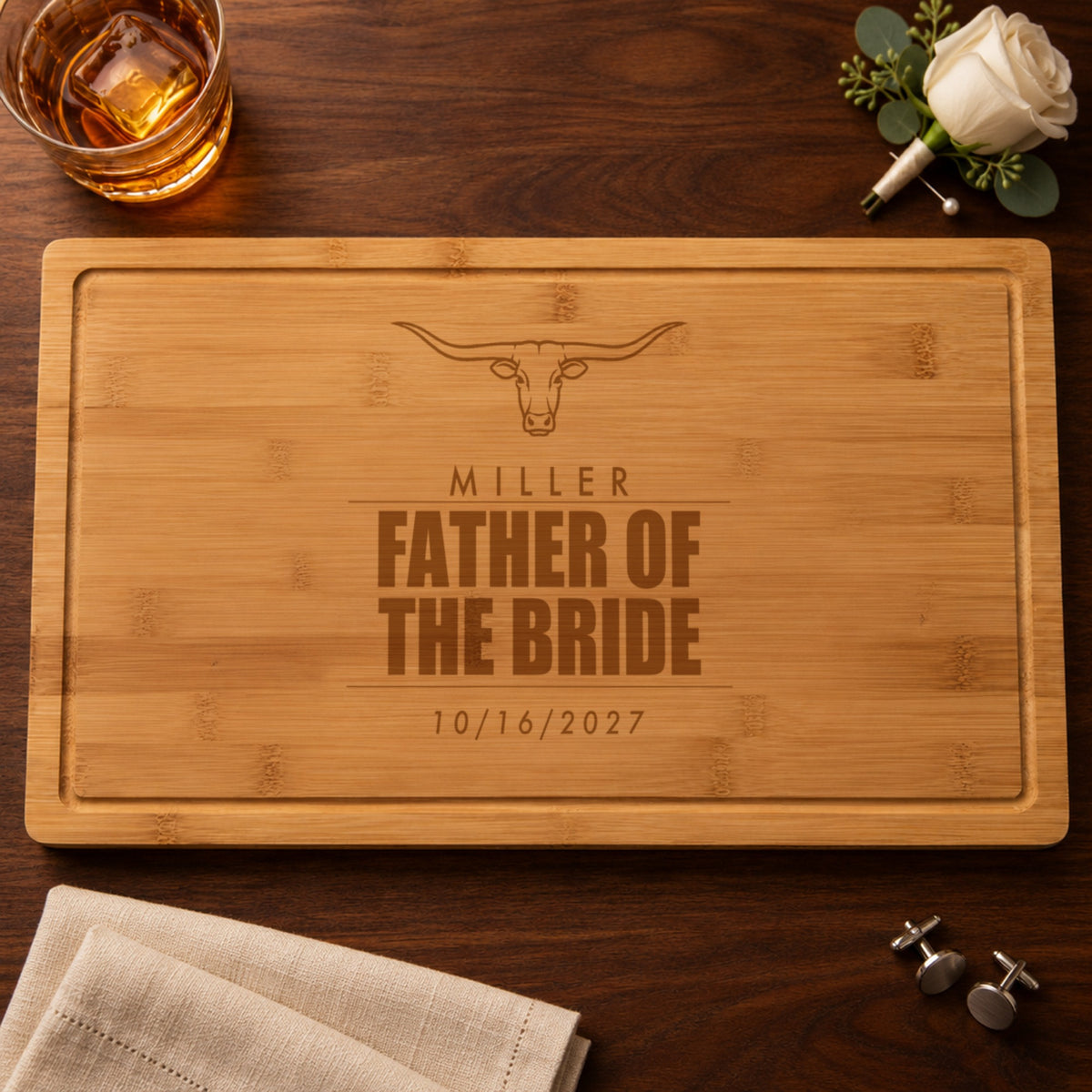 Personalized father of the bride cutting board engraved with a longhorn, MILLER, FATHER OF THE BRIDE and 10/16/2027, beside a white rose boutonniere and whiskey