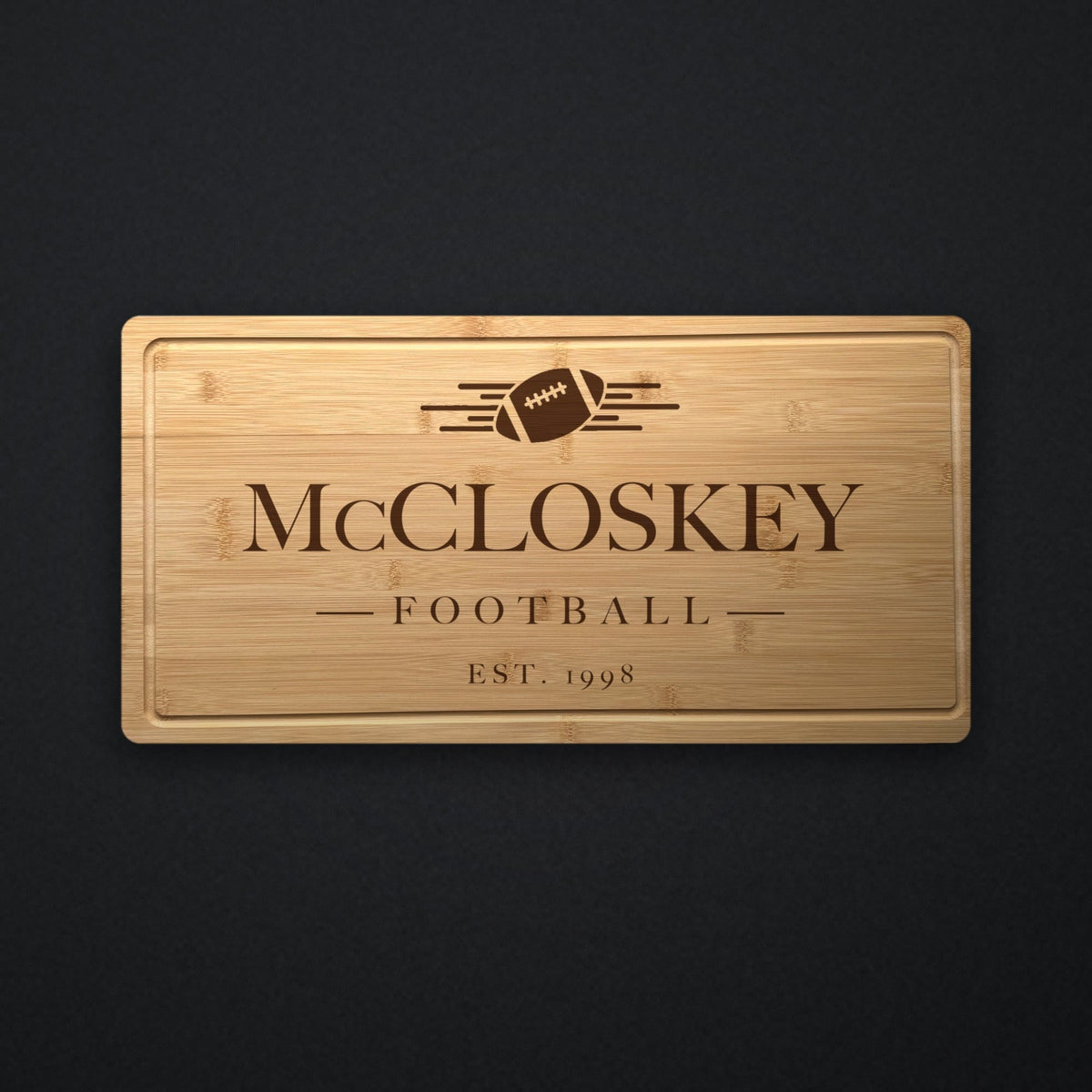 Personalized football tailgate cutting board in solid bamboo on dark slate, engraved with a football and speed lines above the family name and the year.