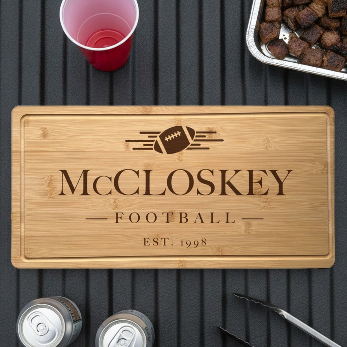 Engraved football bamboo board on the open tailgate of a pickup truck beside a tray of grilled burnt ends, two cans, a red cup and grill tongs.