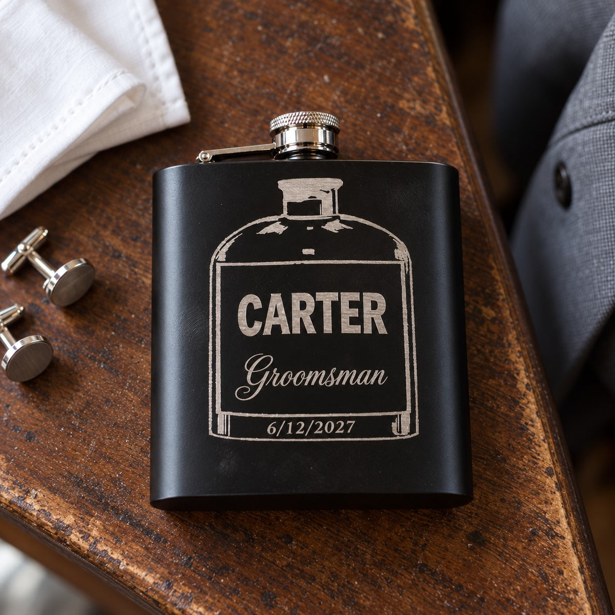 Matte black groomsmen flask engraved with a whiskey bottle design, CARTER, Groomsman in script and the date 6/12/2027, on a dresser beside cufflinks.