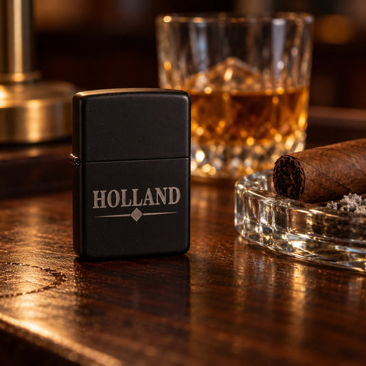 Matte black personalized flip lighter engraved HOLLAND with a diamond line, standing beside a cigar on an ashtray and a glass of whiskey on a walnut bar.