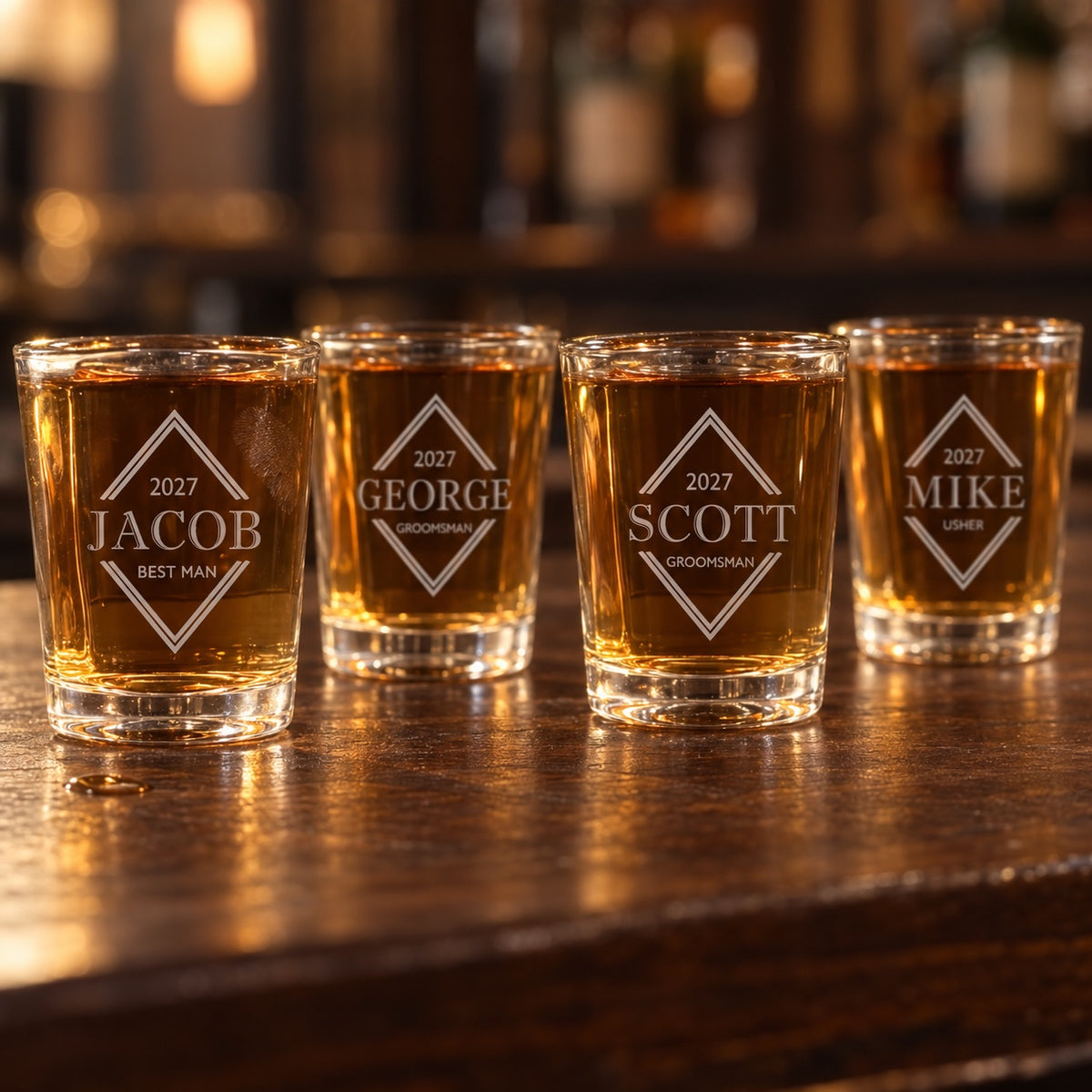 Four personalized groomsmen shot glasses for the wedding party, engraved for a best man, two groomsmen and an usher, lined up on a dark wood bar.