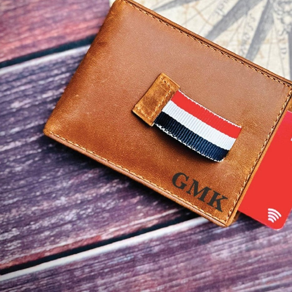 Tan leather bifold wallet engraved GMK in the lower right corner, its red, white and navy ribbon pull tab sliding a red card out, a personalized groomsmen wallet