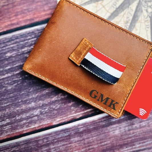 Tan leather bifold wallet engraved GMK in the lower right corner, its red, white and navy ribbon pull tab sliding a red card out, a personalized groomsmen wallet