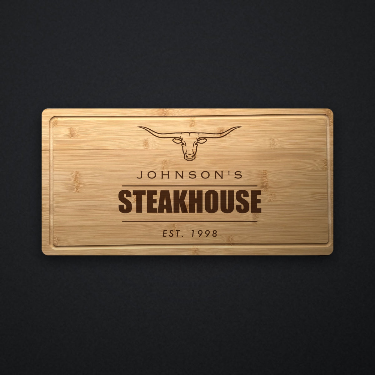 Personalized steakhouse cutting board in solid bamboo on dark slate, engraved with a longhorn steer above the name and the word Steakhouse.