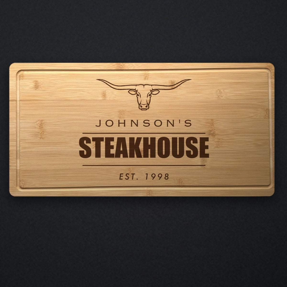 Personalized steakhouse cutting board in solid bamboo on dark slate, engraved with a longhorn steer above the name and the word Steakhouse.