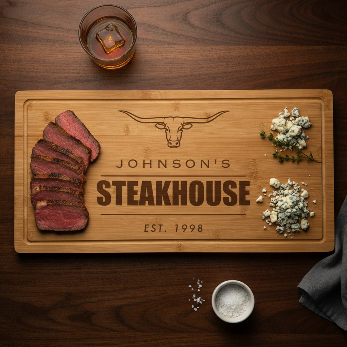 Engraved steakhouse bamboo board on a dark walnut bar top with sliced medium rare ribeye at one end, blue cheese and thyme at the other, beside a bourbon glass.