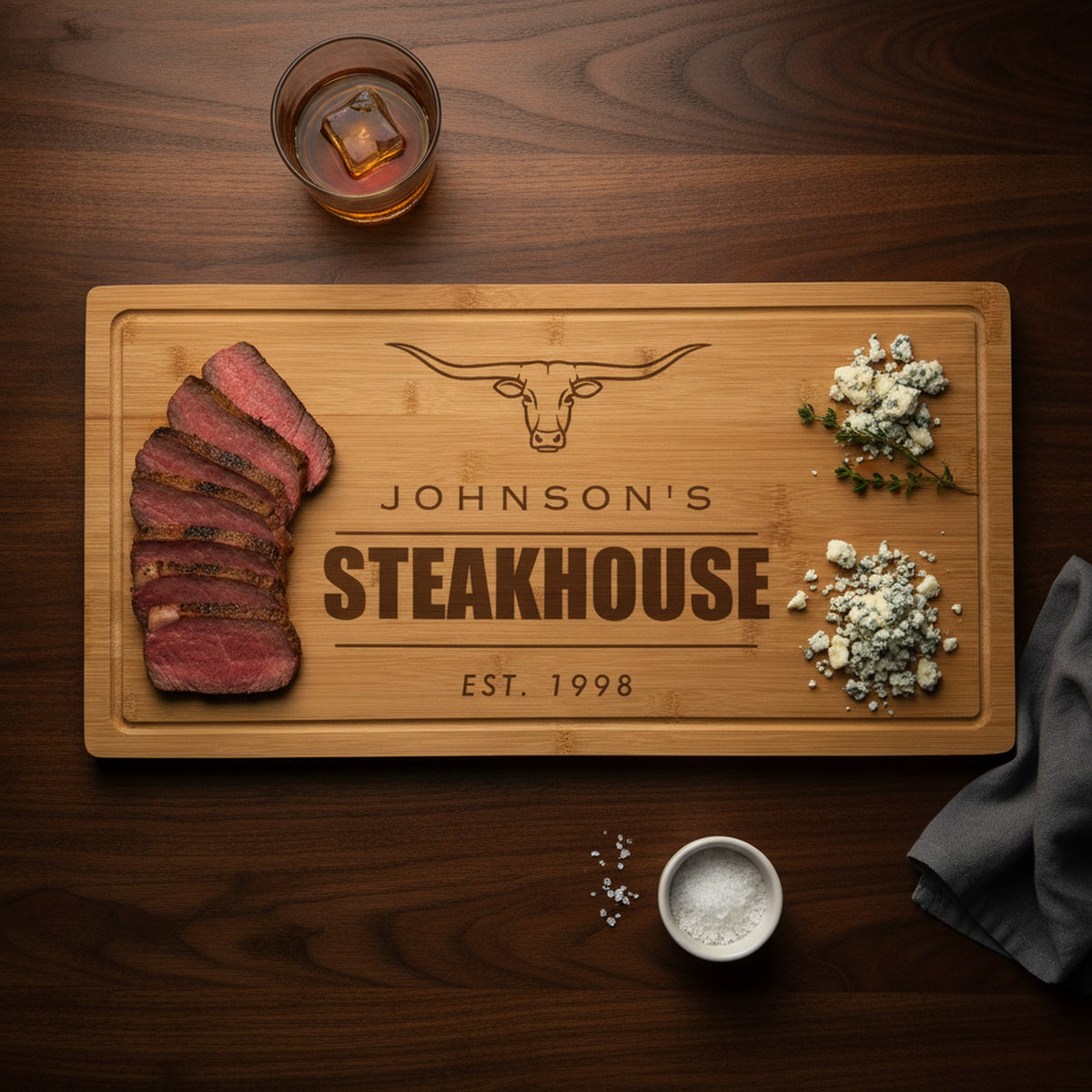 Engraved steakhouse bamboo board on a dark walnut bar top with sliced medium rare ribeye at one end, blue cheese and thyme at the other, beside a bourbon glass.