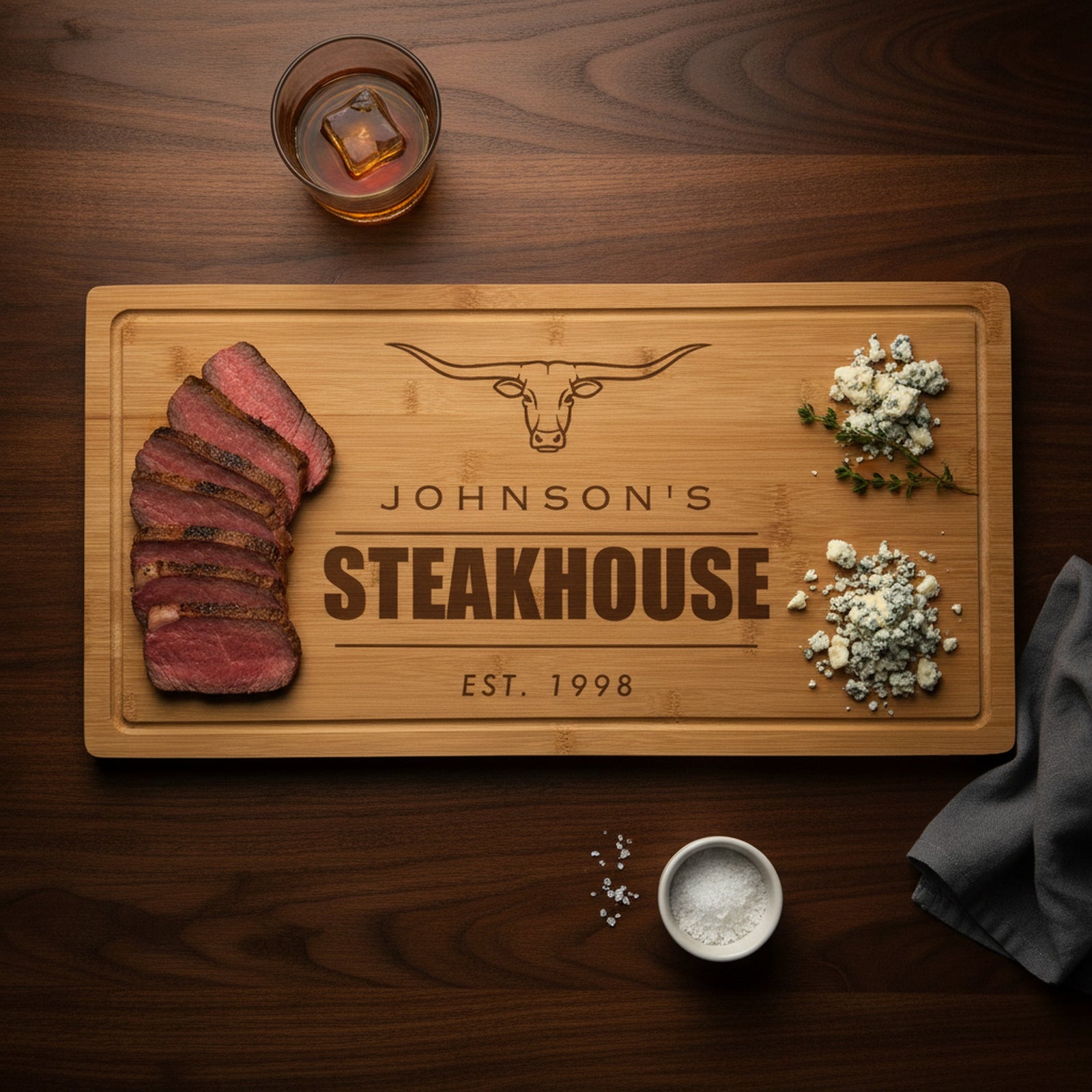 Engraved steakhouse bamboo board on a dark walnut bar top with sliced medium rare ribeye at one end, blue cheese and thyme at the other, beside a bourbon glass.
