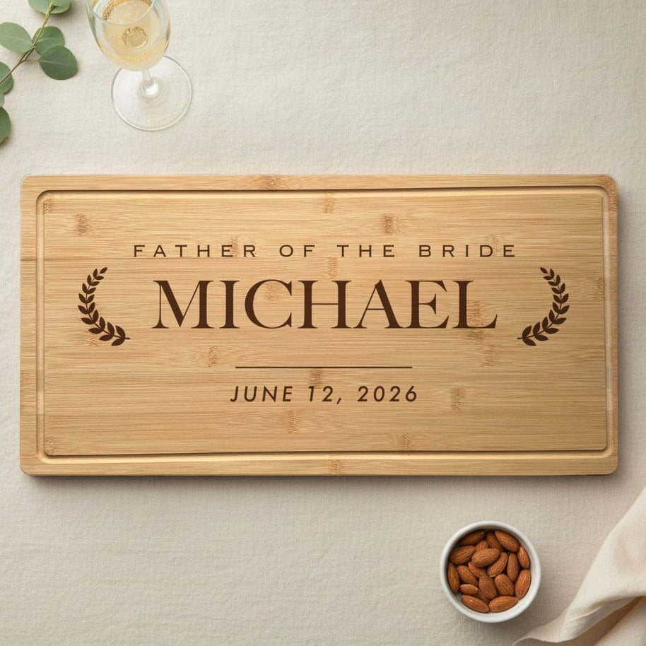 Engraved father of the bride bamboo cutting board laid on an ivory linen table with a champagne glass, eucalyptus sprig and a small dish of almonds beside it.