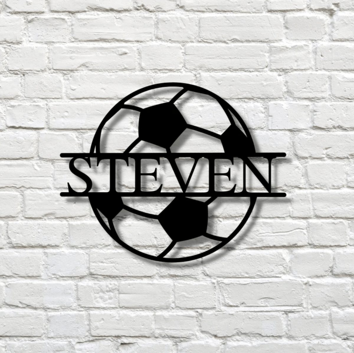 Steel soccer name sign with STEVEN cut across the middle of the ball, mounted on a white painted brick wall, its shadow showing the cutout letters