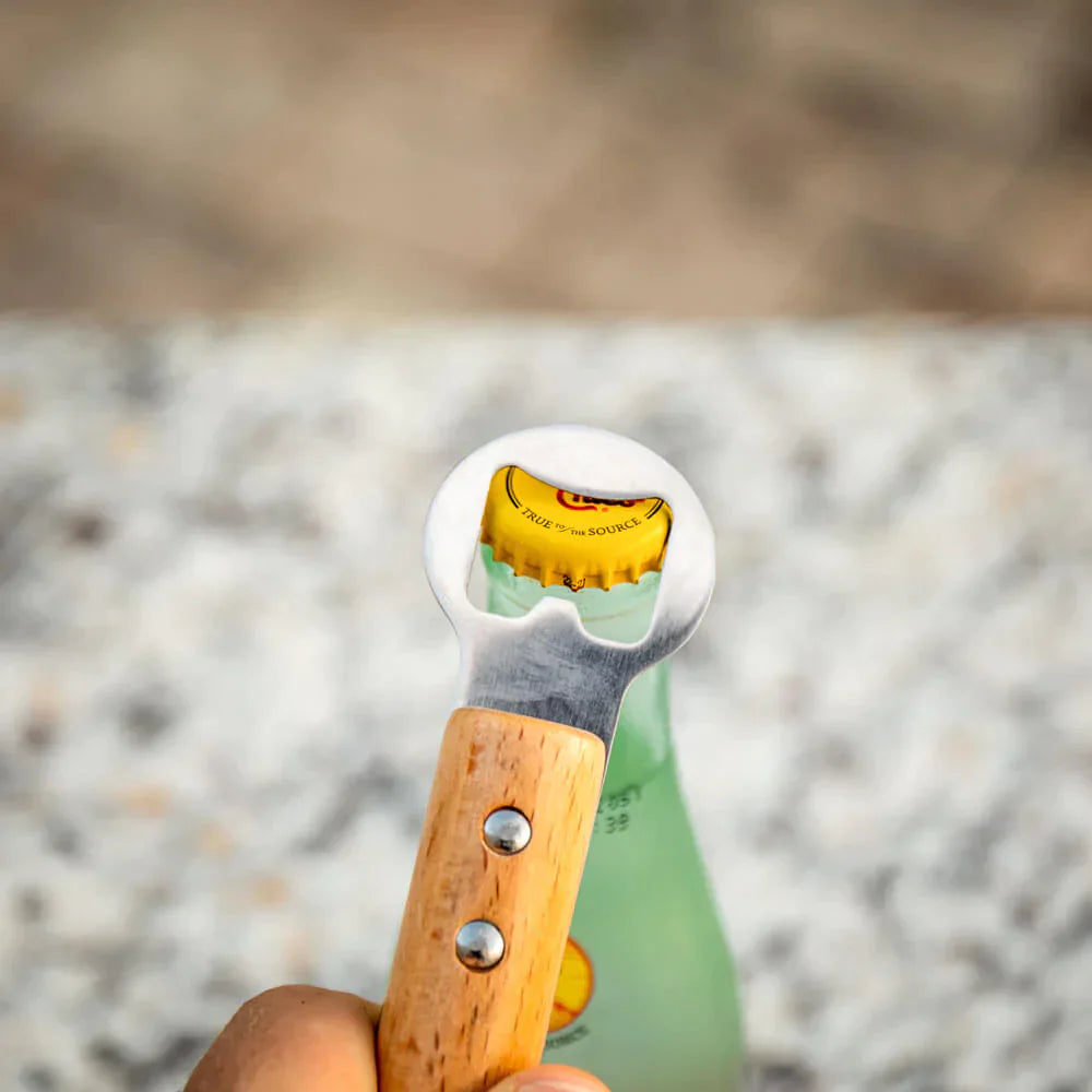 Baseball bat bottle opener with wooden handle and stainless steel head, held upright opening a green bottle against sandy beach background