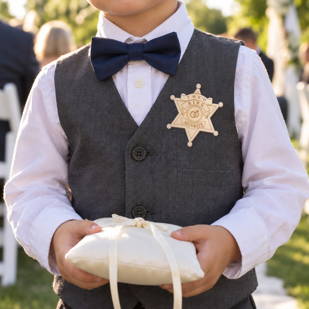 Young ring bearer in a gray vest and navy bow tie wearing a personalized ring security badge engraved THEO while holding a ring pillow