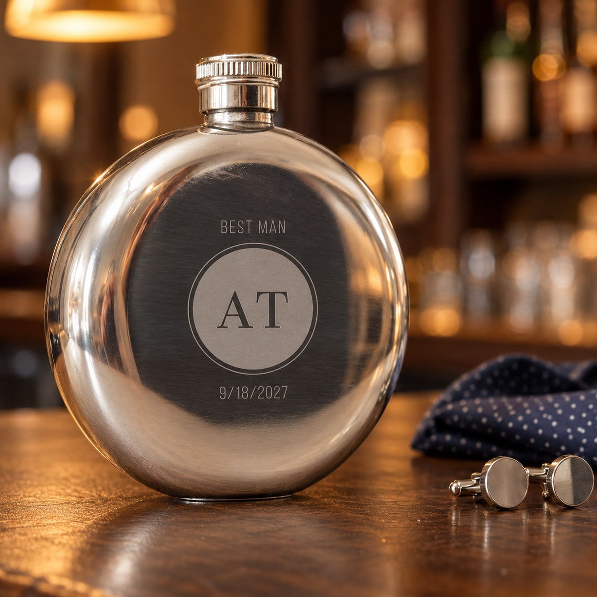 Round polished silver hip flask engraved BEST MAN above a monogram circle with the initials AT and 9/18/2027, a personalized groomsmen flask on a walnut bar.