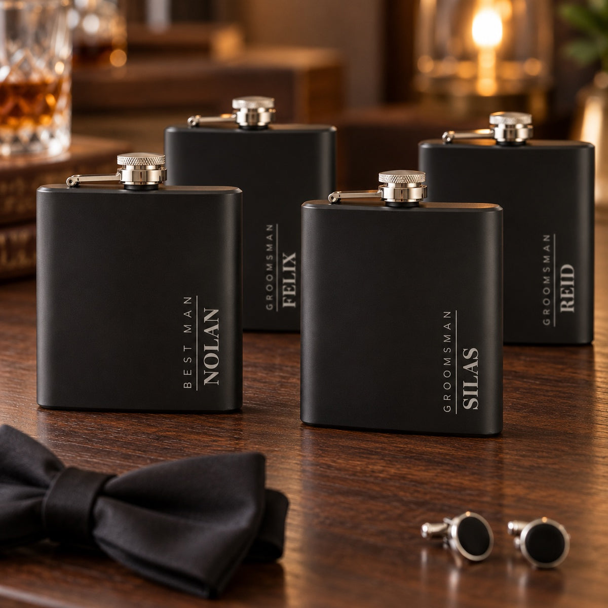 Four engraved flasks for groomsmen on a walnut table with a bow tie and cufflinks: NOLAN as BEST MAN, and SILAS, FELIX and REID each engraved GROOMSMAN