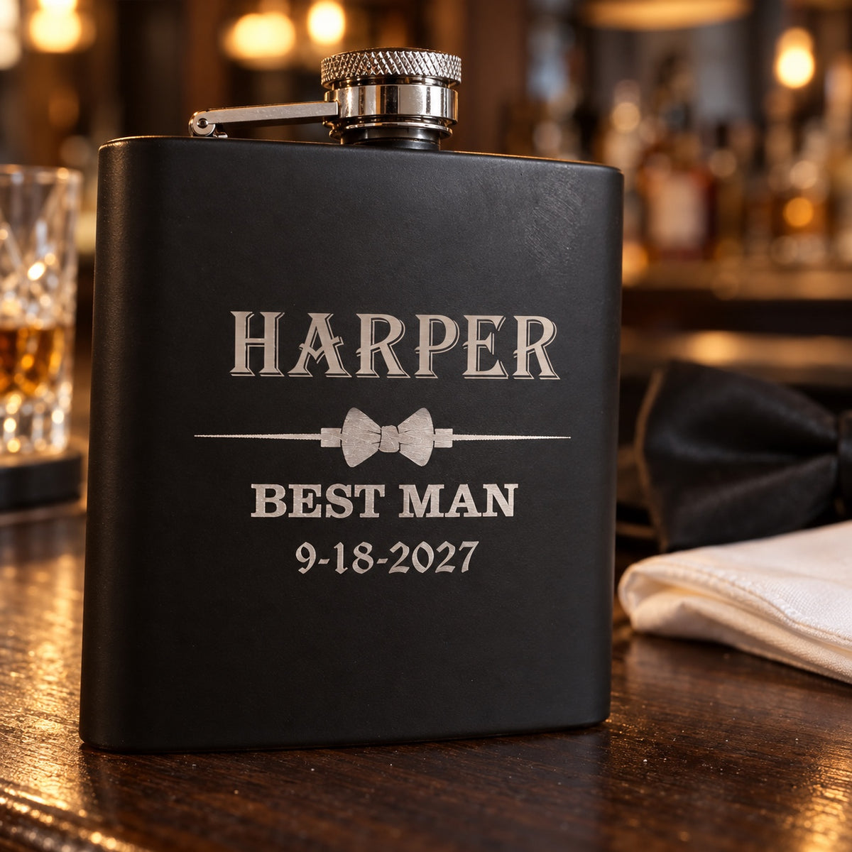 Black groomsmen flask engraved HARPER above a bow tie, BEST MAN and 9-18-2027, a personalized wedding flask standing on a walnut bar beside a bow tie.