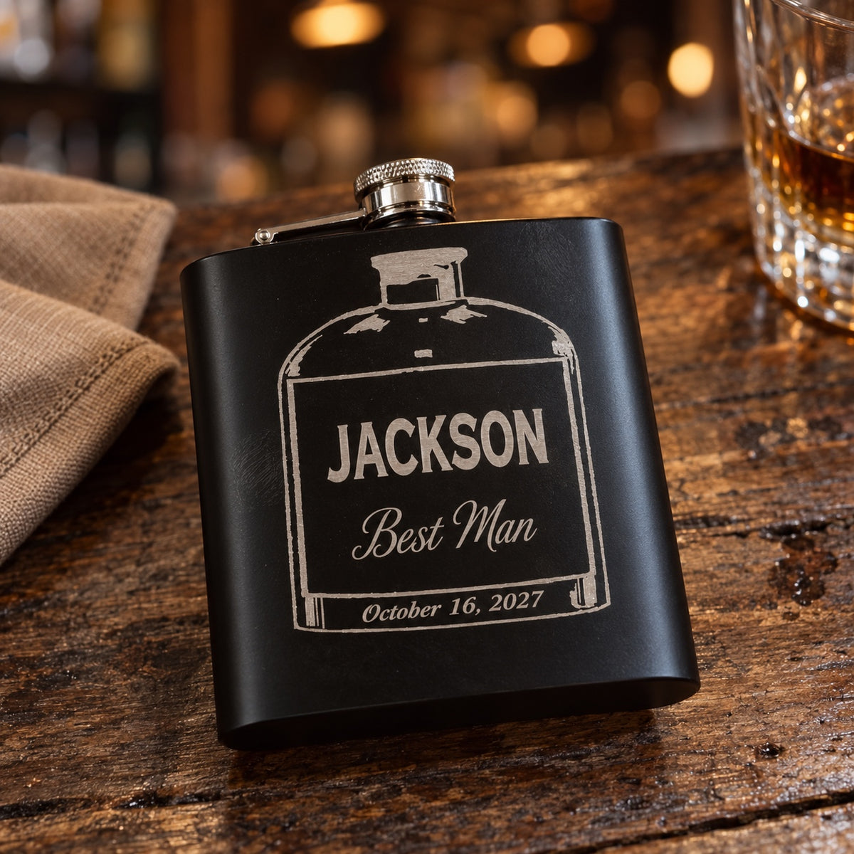 Matte black groomsmen flask engraved with a whiskey bottle outline, JACKSON, Best Man in script and October 16, 2027, a personalized flask on a wood bar.