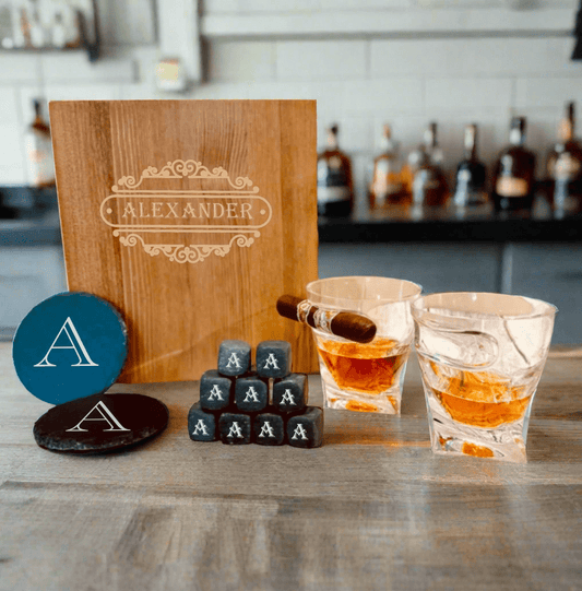 Personalized whiskey glass with built-in cigar rest, six whiskey stones, two slate coasters, and an engraved wooden gift box arranged as a groomsmen gift set.